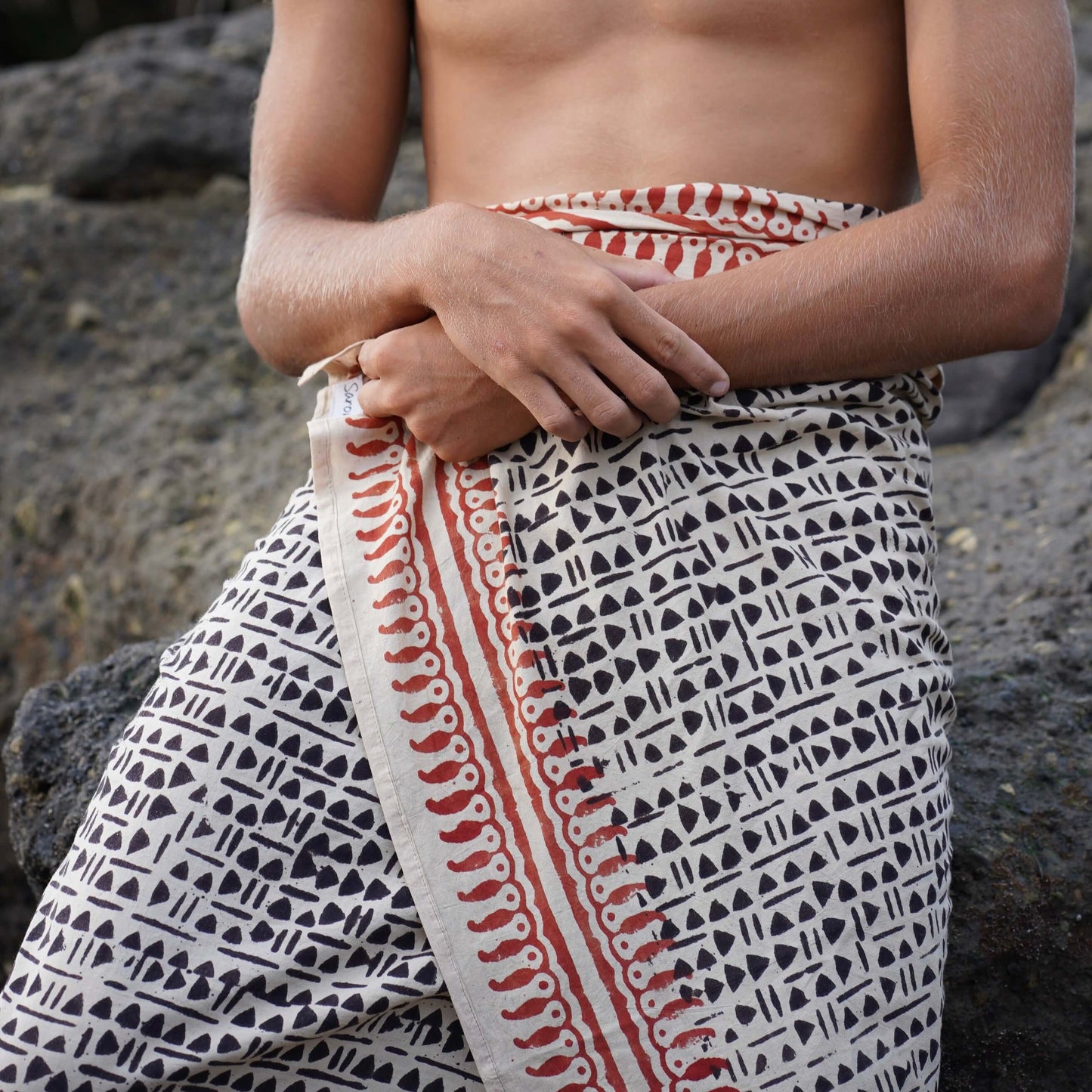 Man sitting outdoors wearing Batik Cotton Mens Sarong in black and brown, styled for casual travel or beachwear.